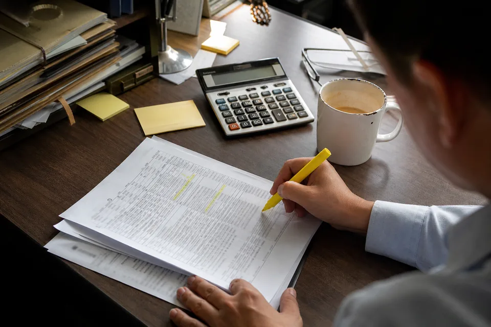 A hand with a highlighter marking up a printed enterprise software order form beside a desk calculator and sticky notes